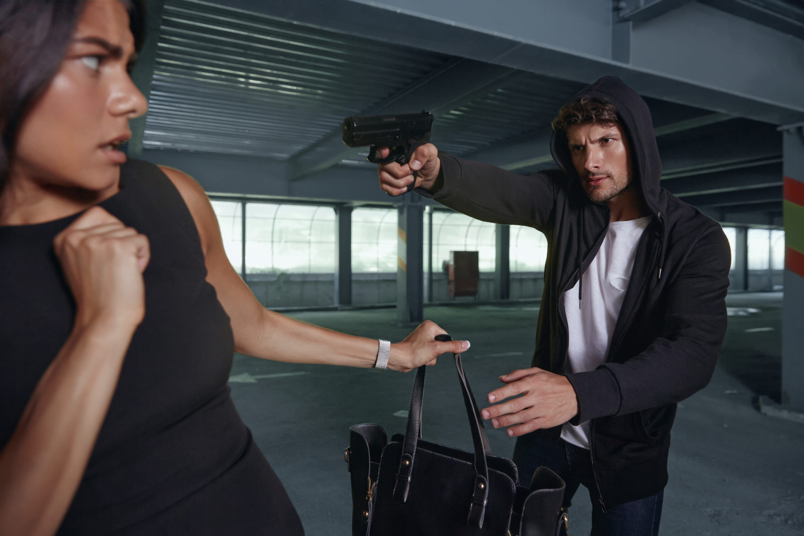 Angry robber want to steal handbag of frightened girl. Male bandit wear black hoodie and hold pistol. Young brunette woman wear black dress. Concept of robbery. Inside parking lot